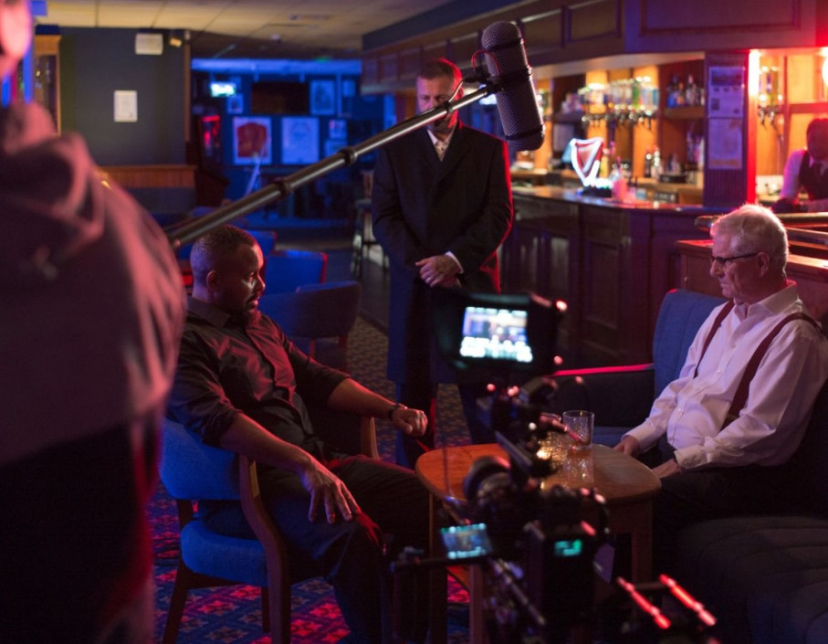 Men seated in. pub setting being filmed with microphones and recording equipment