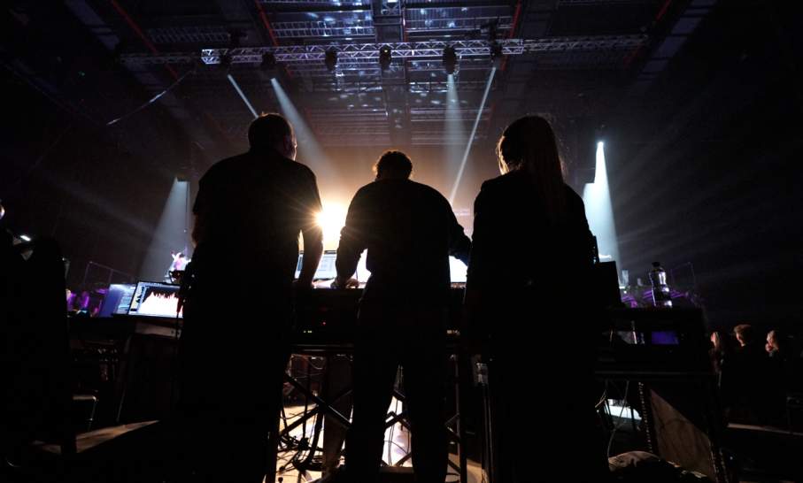 Silhouetted technicians working at a live production control desk in a large indoor venue, with bright stage lights and rigging overhead, as performers appear in the distance on stage.