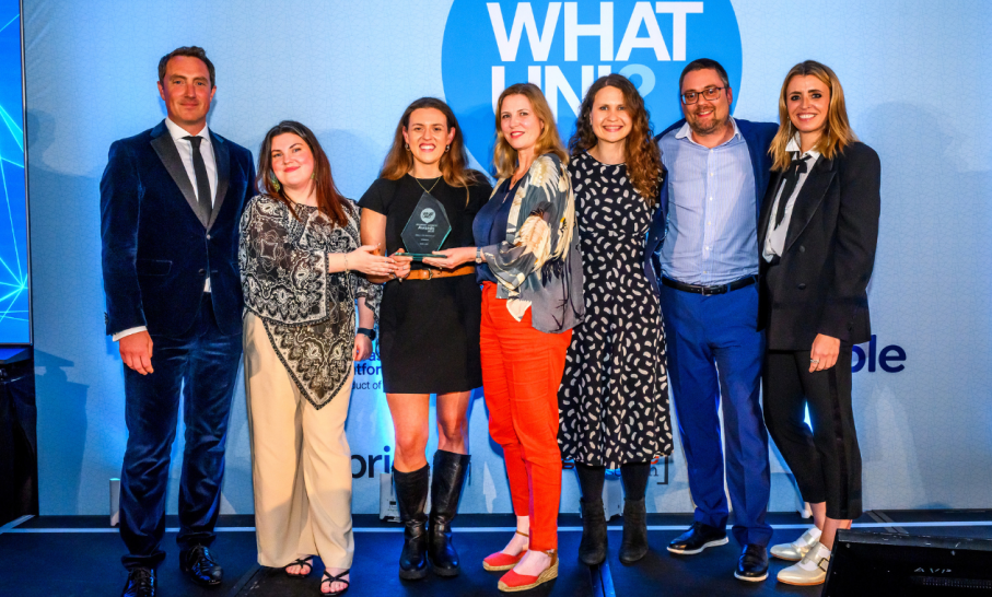 Eight Norland College staff stand together on stage at a Whatuni awards ceremony, with two individuals in the centre holding a glass award trophy. The group is dressed in business attire and positioned in front of a blue backdrop featuring large ‘WHAT UNI’ branding.