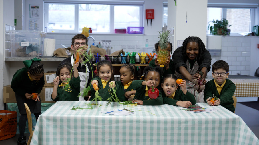 Children and two adults are smiling sitting at a table with green and white checked table cloth, holding carrots up to the camera.
