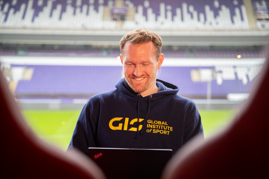 Man wearing branded hoodie sitting at laptop with stadium behind him