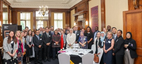 Group photo of around 40 people in business attire standing and smiling in an ornate wood-paneled room with chandeliers and large windows. A round table with papers, cups, and bags is in the foreground, and a banner for Independent Higher Education is visible on the left.
