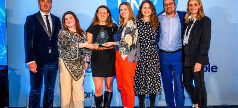 Eight Norland College staff stand together on stage at a Whatuni awards ceremony, with two individuals in the centre holding a glass award trophy. The group is dressed in business attire and positioned in front of a blue backdrop featuring large ‘WHAT UNI’ branding.