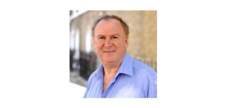 A photo of Tony Millns, an white man with brown greying hair, with a slight smile, wearing a light blue shirt, with a background of light stone buildings.