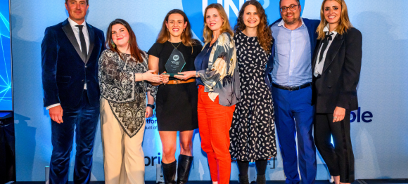 Eight Norland College staff stand together on stage at a Whatuni awards ceremony, with two individuals in the centre holding a glass award trophy. The group is dressed in business attire and positioned in front of a blue backdrop featuring large ‘WHAT UNI’ branding.