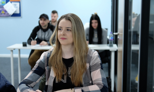 Waltham International College female student wearing a checked shirt and long blonde hair, listening in a lesson at a desk holding a pen, with other students behind her.
