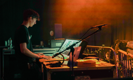 Person operating a digital mixing or lighting control desk backstage at a live event, lit by orange stage smoke, with microphones, laptops and audio equipment visible around the workstation and a performance area beyond.