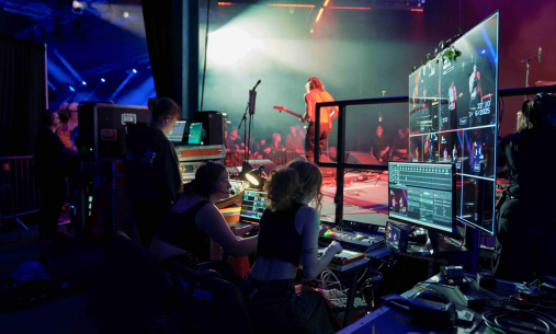 Behind-the-scenes view of a live event, with technicians operating sound and video equipment at a control desk while a musician performs on stage under bright coloured lights, and multiple screens display live camera feeds and production software.