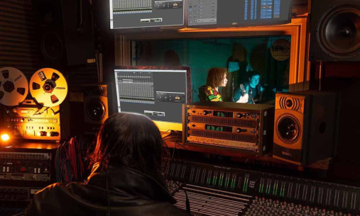 A recording studio with someone recording at a screen, and someone singing at a microphone behind a window