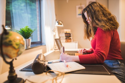 A person with long brown hair wearing a red jumper studying at a desk in a bright room