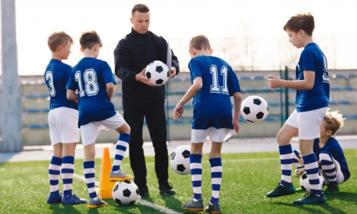 Four boys in blue and white football shirts with black and white footballs being coached by a man in black sportswear