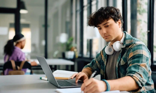 Male student at desk with laptop making notes