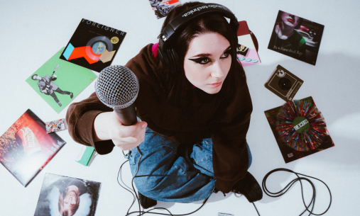 Person sitting on floor surrounded by records wearing headphones and holding up a microphone
