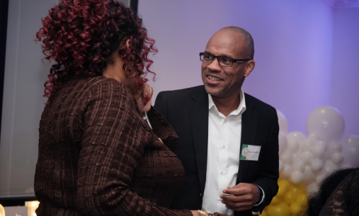 Two people conversing at an event with white and yellow balloons in the background; one in a brown jacket, the other in a black suit with a blurred face.