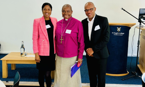Three individuals in formal attire at St Hugh's College, Oxford; one in a pink blazer, one in clerical clothing with a cross necklace, and one in a black suit.